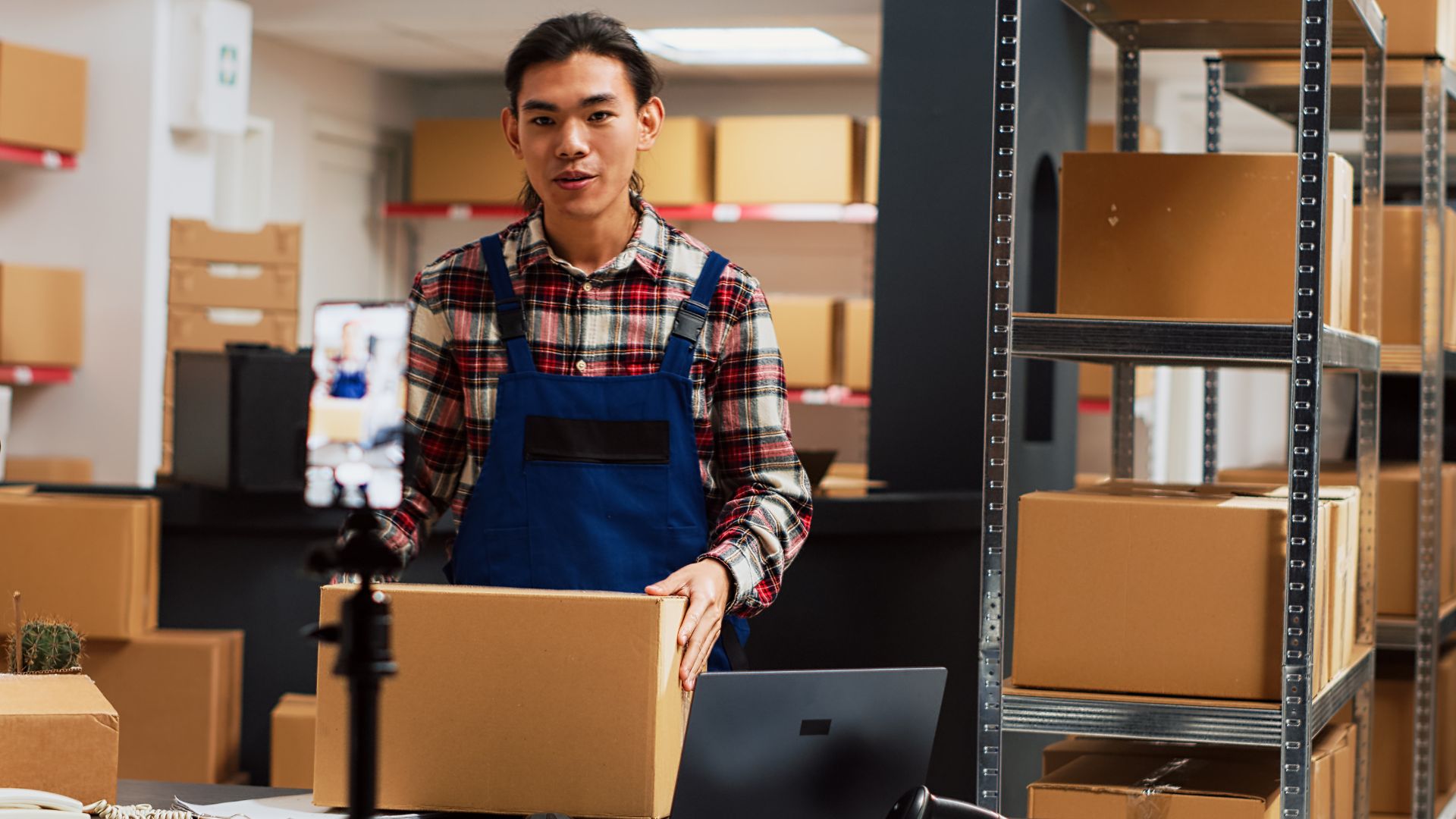 Warehouse worker in plaid shirt and blue apron standing with cardboard boxes