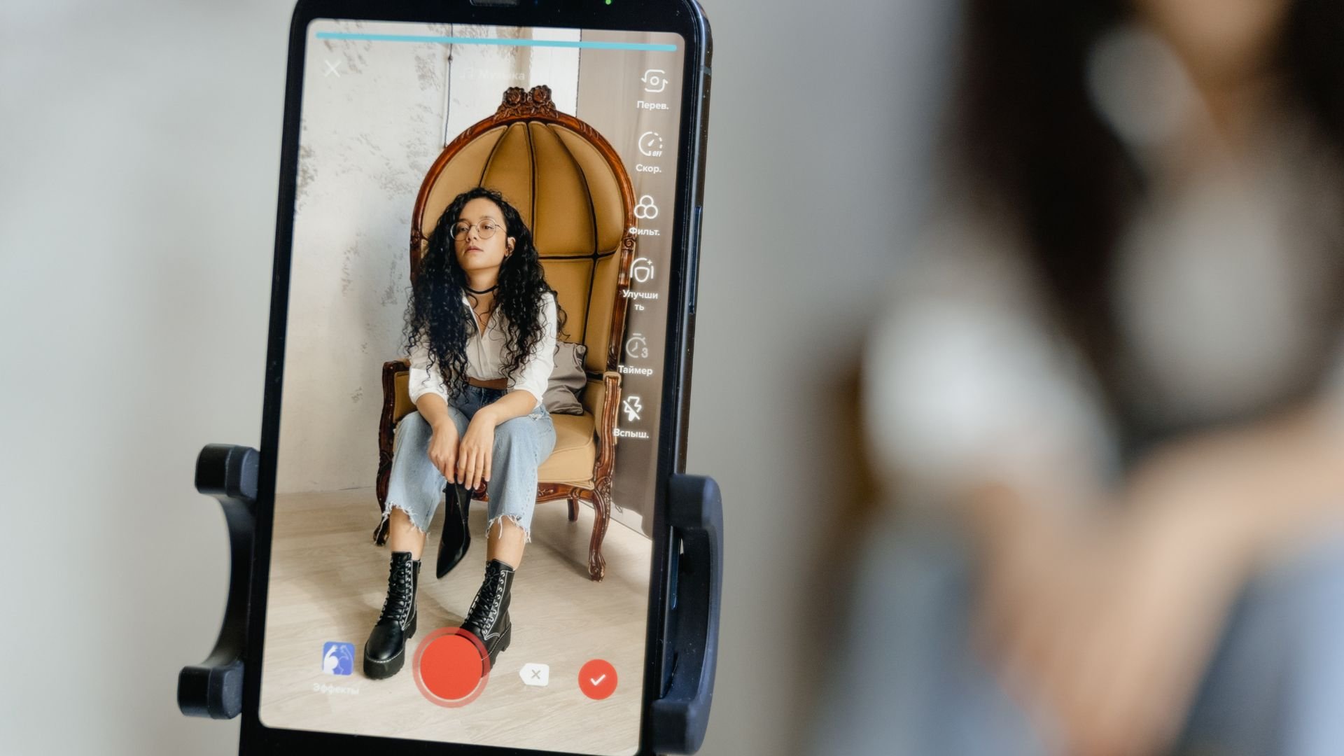 Person with curly hair sitting in ornate chair, viewed through smartphone screen